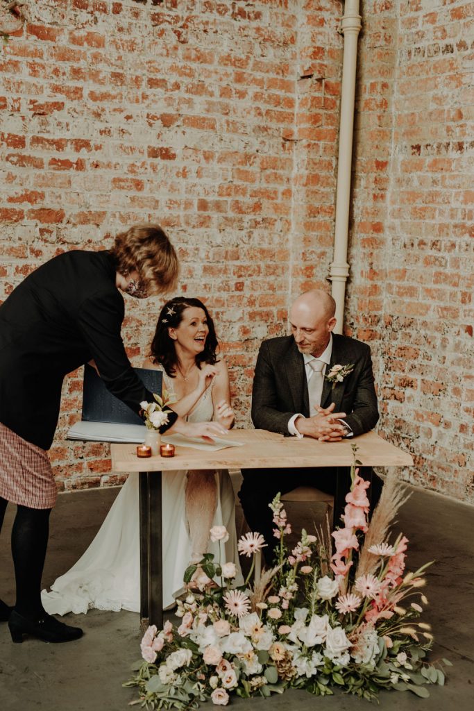 Bride & Groom signing marriage certificate. Floral Ground arrangement sits in front of registry table
