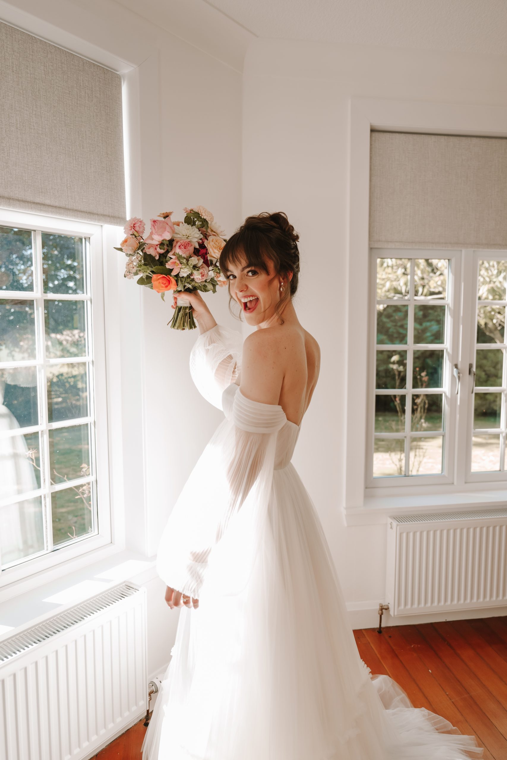 Wedding Flowers and florals -Bride in an off-the-shoulder white wedding dress smiles excitedly while holding up a colourful bouquet by a sunlit window in a bright room.

