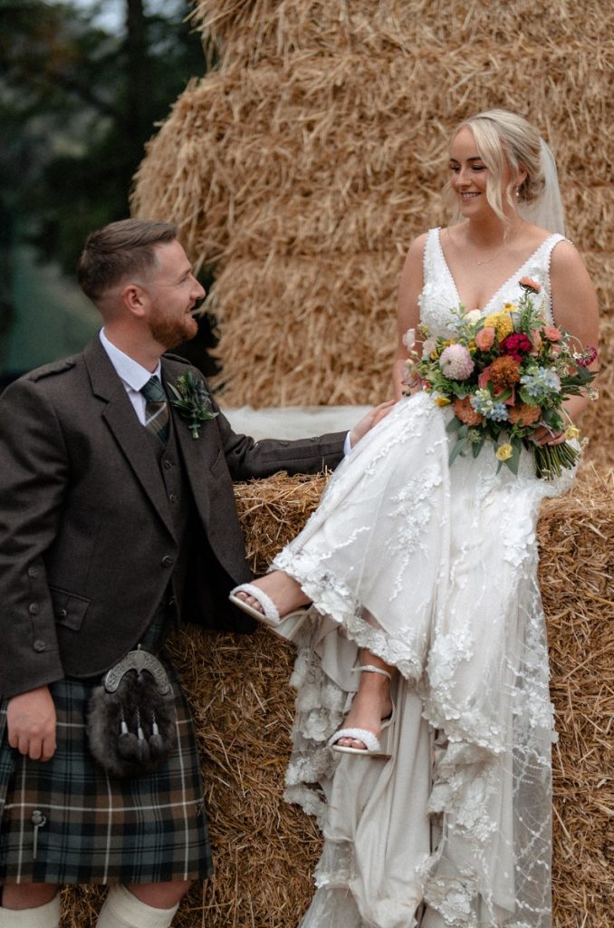 Bride in a white lace wedding gown sits on a hay bale, holding a colorful bouquet as her groom in a kilt smiles at her nearby.