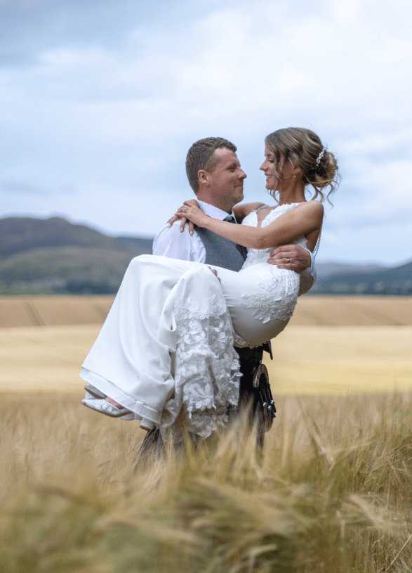 Bride being carried by groom in a white wedding dress and suit, smiling in a grassy field with hills and blue sky in the background.