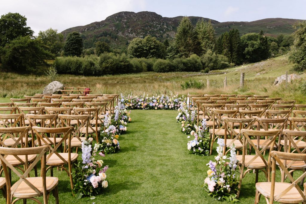 cardney-steadings-wedding-florist-perthshire 2 - Fraoch Floral Design Outdoor wedding ceremony setup on a grassy field with wooden chairs and a flower-lined aisle, mountains in the distance.