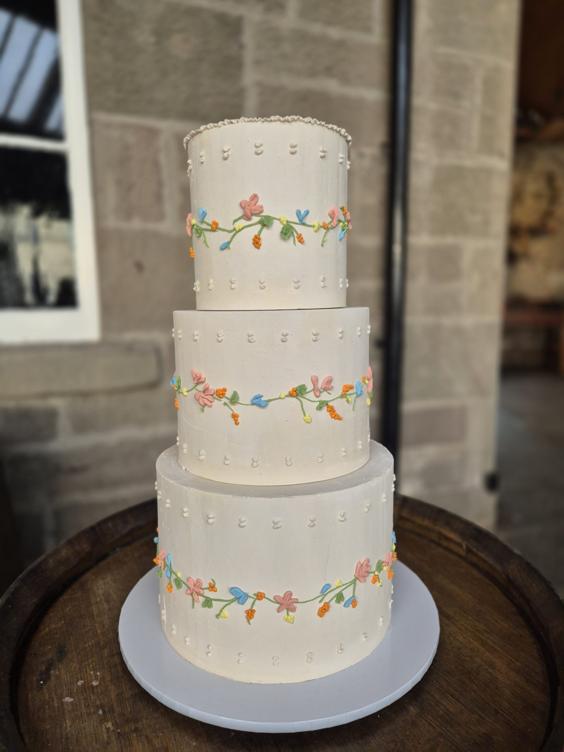 Tall white multi-tier cake with pastel flower garlands and dot borders, set outdoors on a round stand.