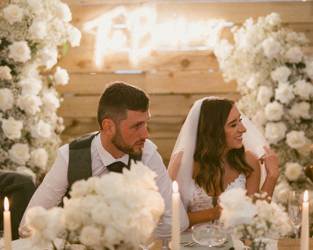 Bride and groom sit at a candlelit wedding reception table flanked by white flowers, smiling at each other.
