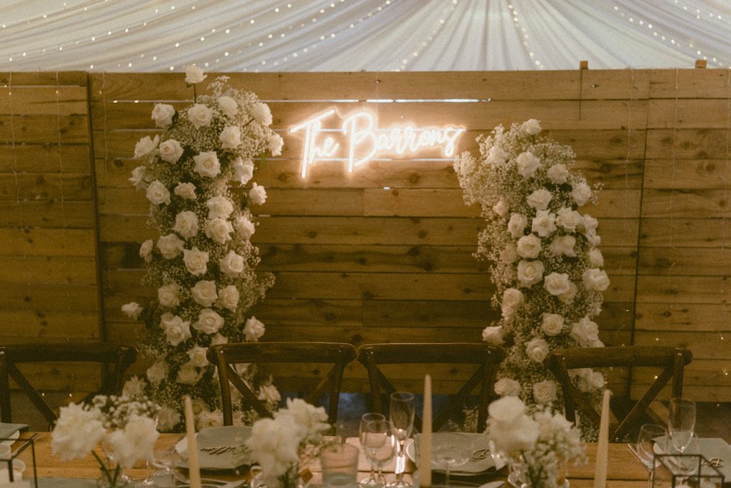 Wooden barn-style reception with two tall white-flower arches framing a neon'The Barn' sign above a long dining table