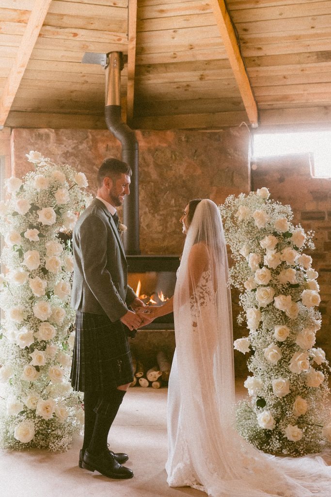 Bride and groom hold hands facing each other between tall white rose arches in a rustic indoor wedding by a fireplace.