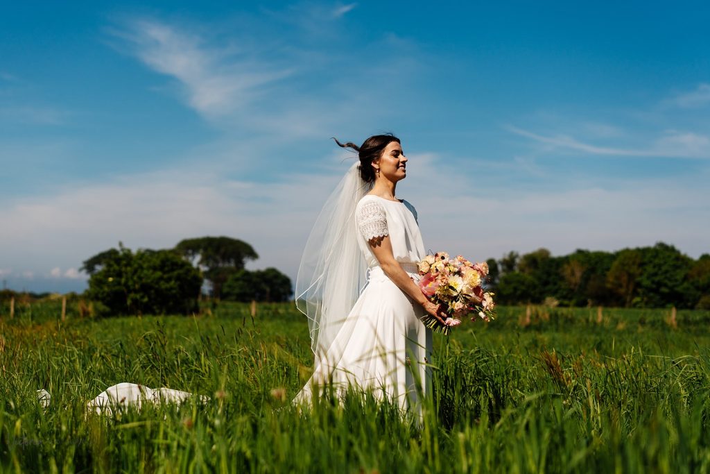 falside-mill-wedding-florist-fife - Fraoch Floral Design Fife Wedding Flowers. Bride in a white wedding dress with veil, holding a bouquet, standing in a green field under a clear blue sky.