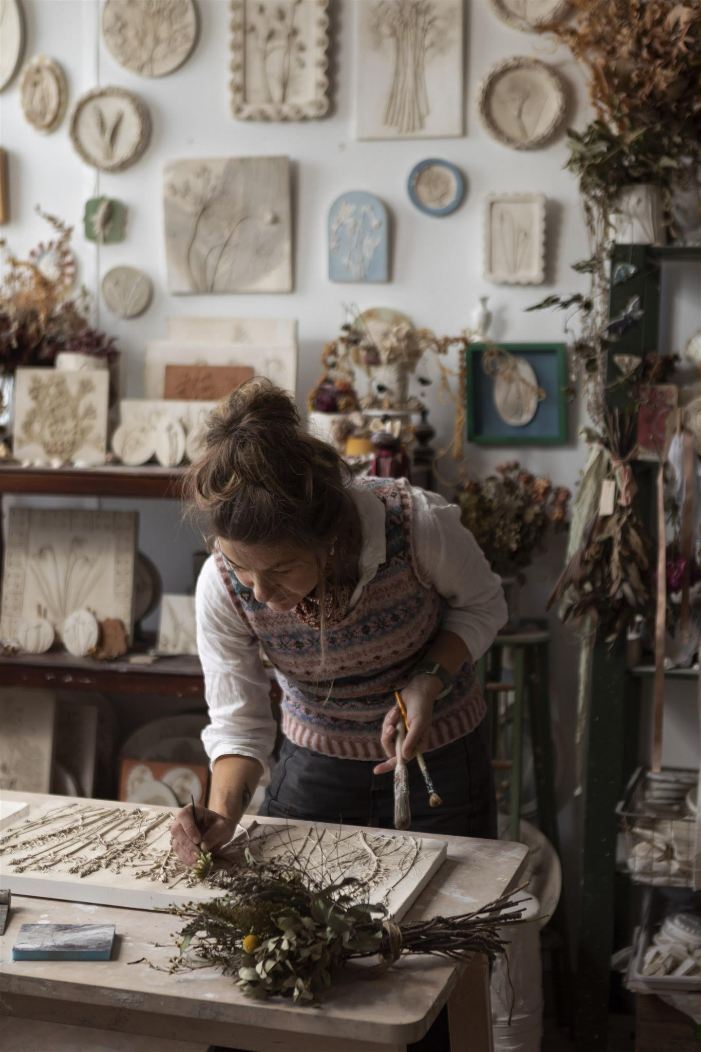 imprint-casts-fife - Fraoch Floral Design Woman arranging dried botanicals on a table in a craft studio with wall-mounted art pieces behind her.