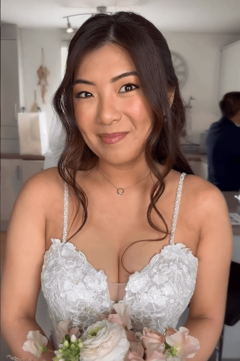jennifer mclauchlan mua - Fraoch Floral Design Woman in a lace bridal top holding a pastel flower bouquet, smiling warmly in a bright room.