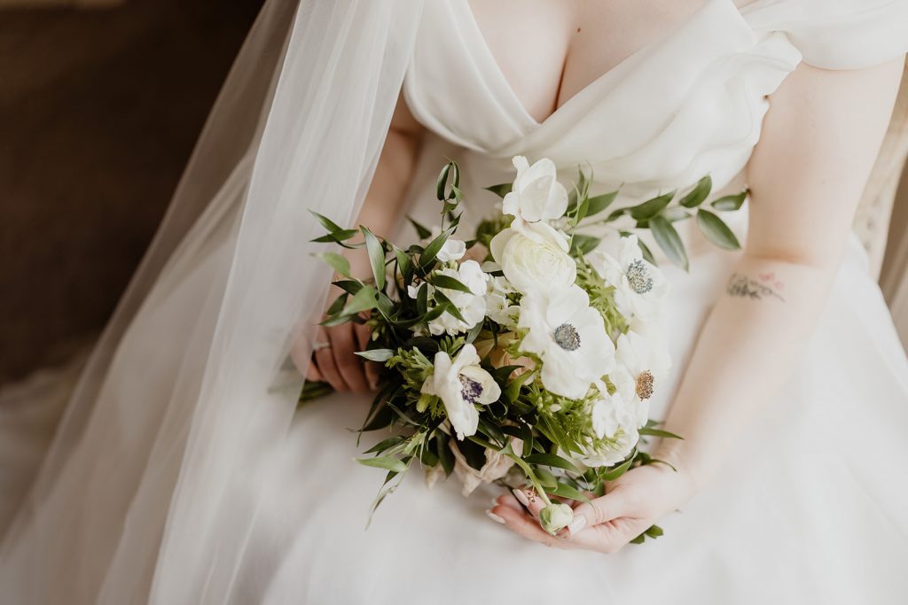 Bride in a white wedding dress holding a white bouquet with greenery and veil in the background