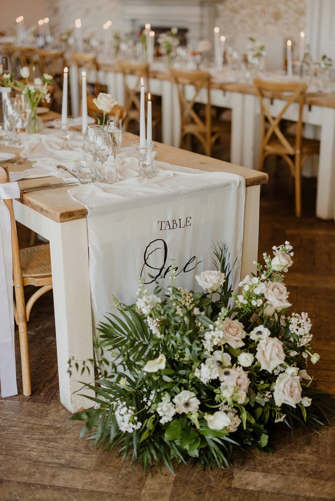 Wedding reception table with a white cloth runner, tall candles, glassware, and a lush floral arrangement at the table end.