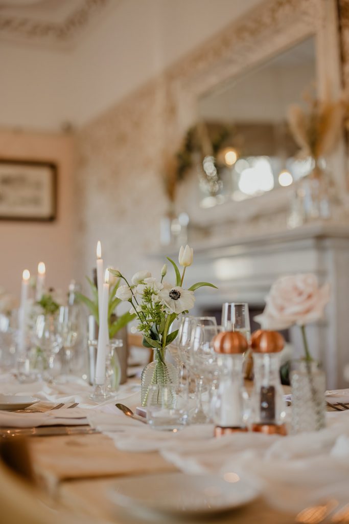 Elegant dining table set with white flower centerpiece, candles, and glassware in a softly lit room.