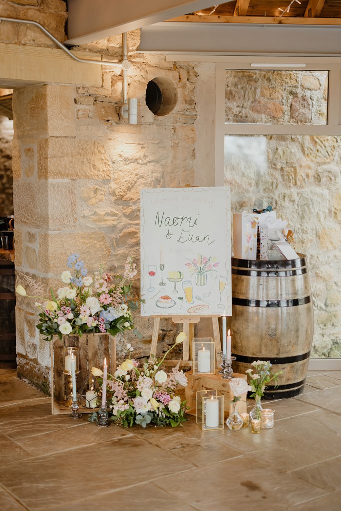 Wedding welcome board on an easel beside a barrel, surrounded by flowers and candles in a stone-walled venue.