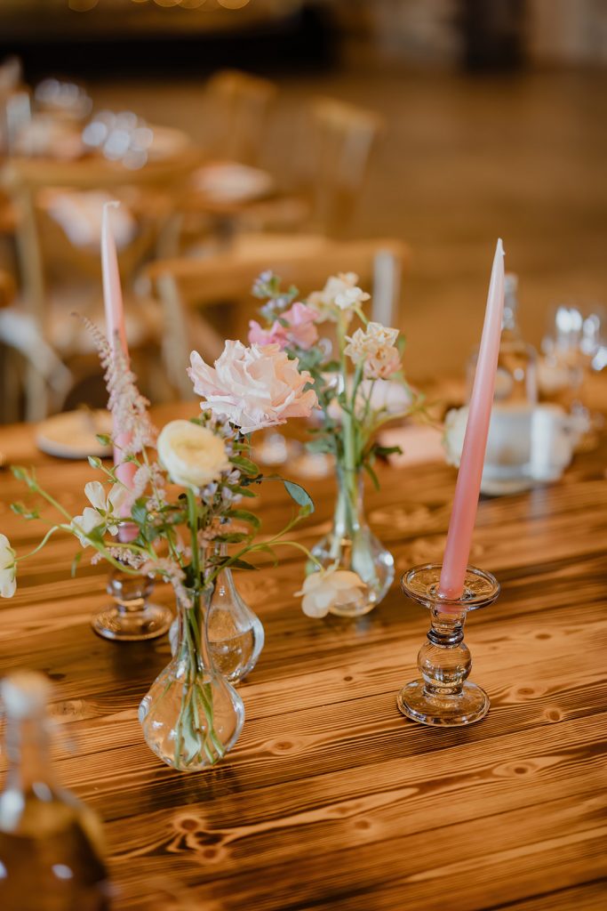 Table decor with pink taper candles in glass holders and pastel floral arrangements in small vases on a wooden table.