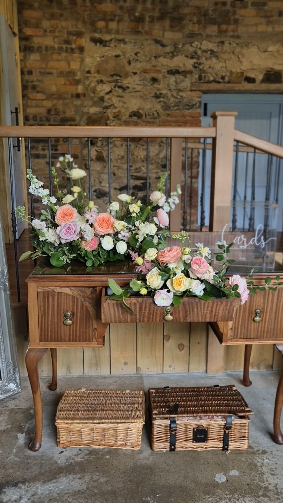 ROSSIE-ON-EARN-WEDDING-FRAOCH-FLORAL-DESIGN 11 - Fraoch Floral Design Rustic wooden dresser with a pastel floral arrangement of roses and greenery against a brick wall.