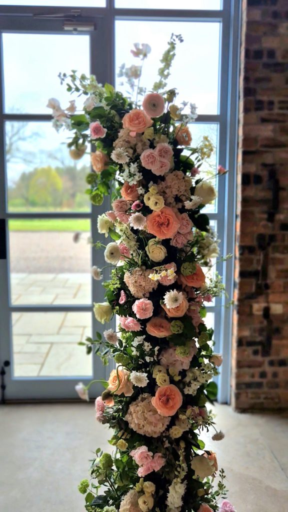 ROSSIE-ON-EARN-WEDDING-FRAOCH-FLORAL-DESIGN 15 - Fraoch Floral Design Tall vertical floral arrangement of pastel pink, peach, and white roses with white blossoms and greenery, set indoors by a large window.