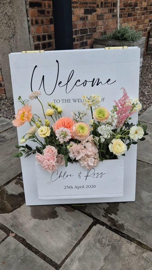 ROSSIE-ON-EARN-WEDDING-FRAOCH-FLORAL-DESIGN 6 - Fraoch Floral Design White wooden welcome sign with pastel flower arrangement in front; text reads'Welcome to the wedding of Chloe & Ross' and '25th April 2026'.