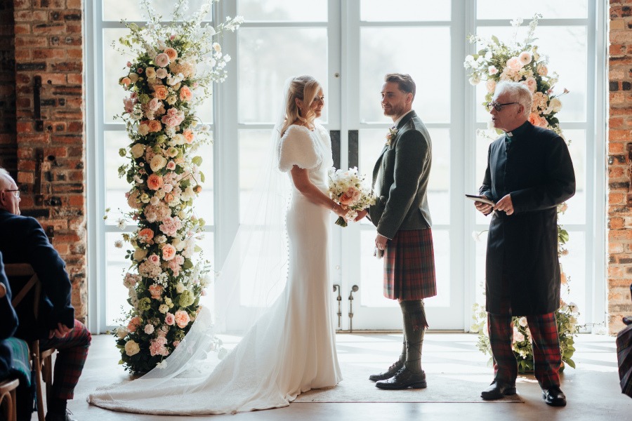 Rossie-on-the-earn-perthshire-wedding-fraoch-floral-design 10 - Fraoch Floral Design Bride in a white gown and veil holds a bouquet, facing her groom in a tartan kilt as an officiant presides by floral arches near a bright window.