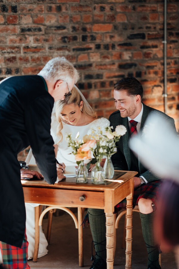 Rossie-on-the-earn-perthshire-wedding-fraoch-floral-design 11 - Fraoch Floral Design Bride and groom signing a document at a wooden table during their wedding, assisted by an older man, with a bouquet in front and a brick wall backdrop.
