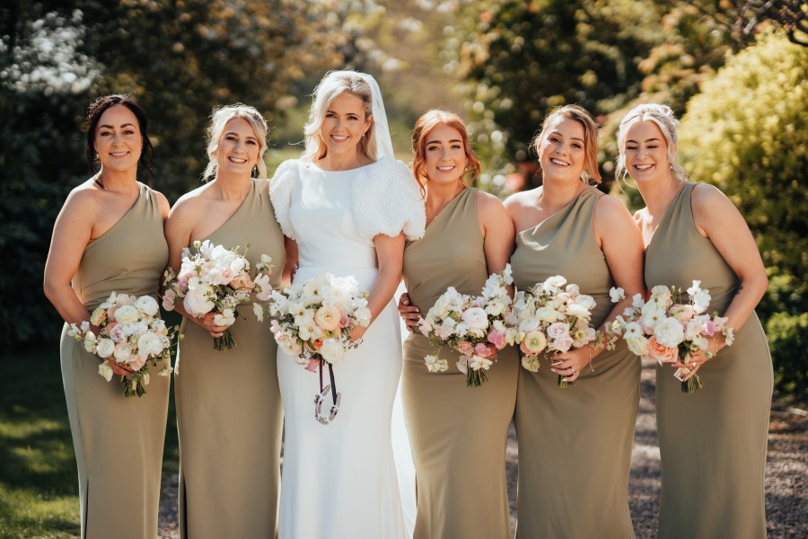 Rossie-on-the-earn-perthshire-wedding-fraoch-floral-design 15 - Fraoch Floral Design Bride in a white wedding dress with fur sleeves stands center with five bridesmaids in sage gowns outdoors, each holding pastel bouquets.