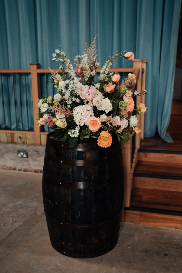 Rossie-on-the-earn-perthshire-wedding-fraoch-floral-design 2 - Fraoch Floral Design Pastel flower arrangement in a dark wooden barrel on a concrete floor near a wooden staircase with blue drapes in the background.