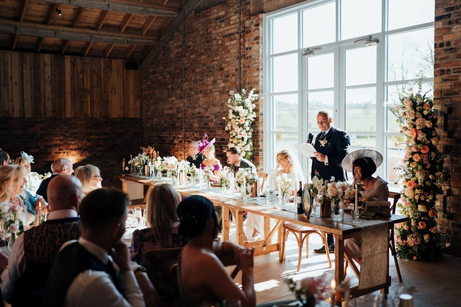 Rossie-on-the-earn-perthshire-wedding-fraoch-floral-design 22 - Fraoch Floral Design Wedding reception in a brick barn: guests sit at a long wooden table as a man in a suit gives a toast by the windowed wall with floral centerpieces.