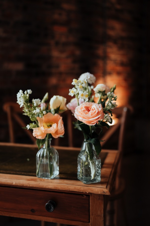 Rossie-on-the-earn-perthshire-wedding-fraoch-floral-design 4 - Fraoch Floral Design Two small glass vases holding peach and white flowers on a rustic wooden table, brick wall backdrop behind them