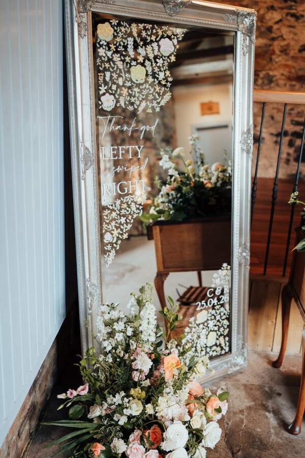 Rossie-on-the-earn-perthshire-wedding-fraoch-floral-design 5 - Fraoch Floral Design Ornate silver-framed mirror with floral decals and a flower arrangement at its base; a wedding sign on the mirror reads “Thank you,” with the words “LEFTY swiped RIGHT” and the couple’s initials and date in the corner.