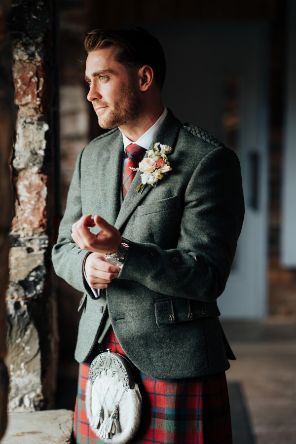 Rossie-on-the-earn-perthshire-wedding-fraoch-floral-design 6 - Fraoch Floral Design Groom in a green tweed jacket and red plaid kilt with a boutonniere, adjusting his watch by a stone wall.