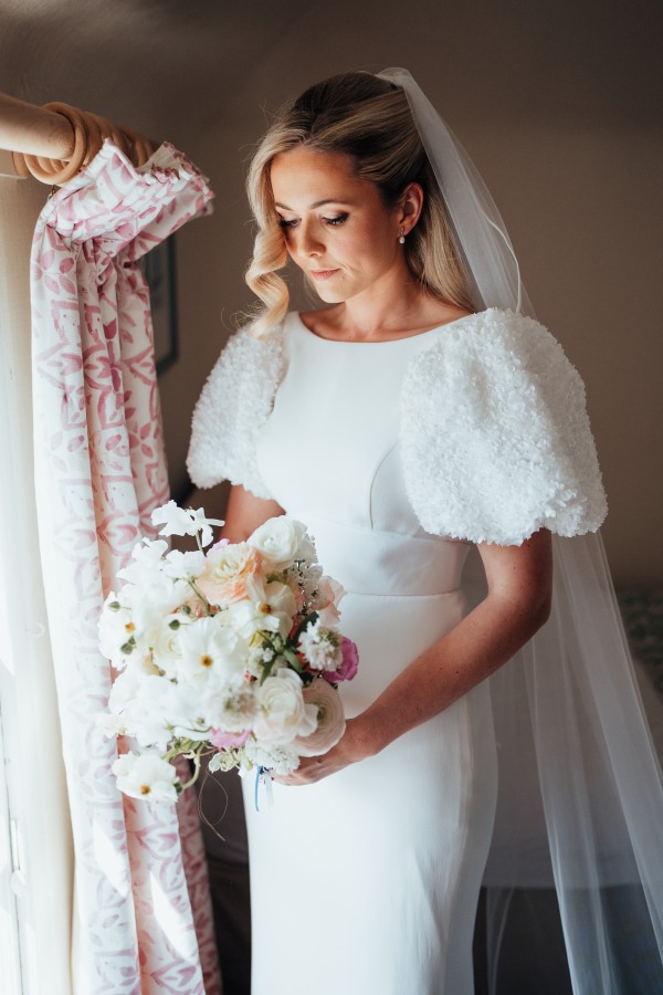 Rossie-on-the-earn-perthshire-wedding-fraoch-floral-design 7 - Fraoch Floral Design Bride in a white wedding gown with fluffy puff sleeves, holding a ivory and pastel bouquet by a window while looking down gracefully.