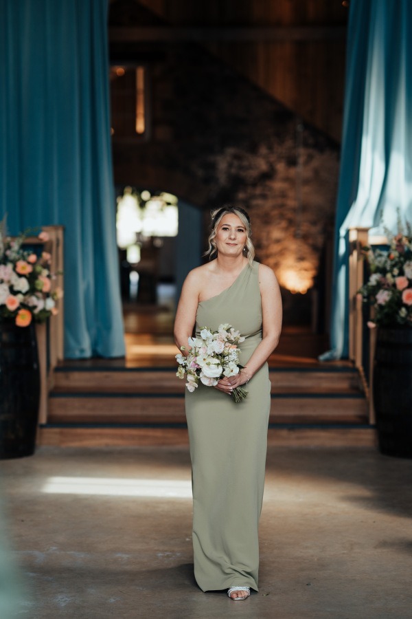 Rossie-on-the-earn-perthshire-wedding-fraoch-floral-design 9 - Fraoch Floral Design A woman in a sage green one-shoulder gown holds a bouquet, standing in a decorated wedding venue with blue drapes and warm lighting.