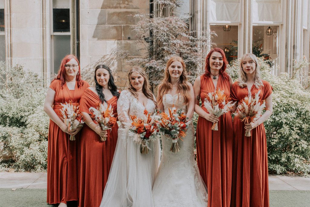 Group of six women posing outdoors: two brides in white gowns flanked by four bridesmaids in red dresses, each holding bouquets.
