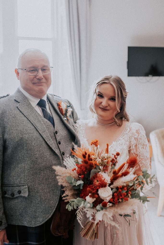 Bride in lace wedding dress holds a vibrant orange bouquet beside an older man in a grey tweed suit and glasses indoors.