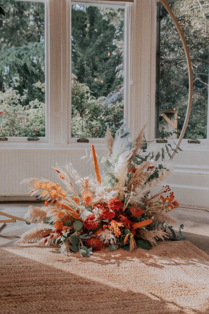Autumn bouquet with orange flowers, dried grasses, and eucalyptus arranged on a woven rug by a large window.