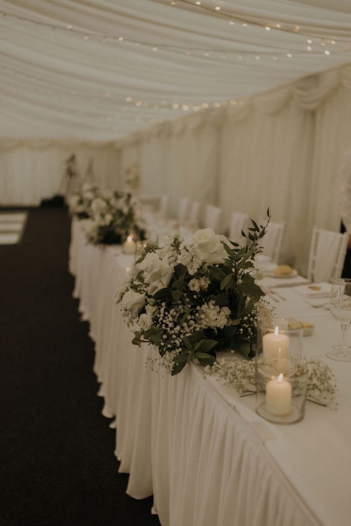 angus-wedding-florist-kinnettles-castle 10 - Fraoch Floral Design Banquet table with a lush white floral centerpiece, candles, and draped white fabric in a softly lit tent setting.