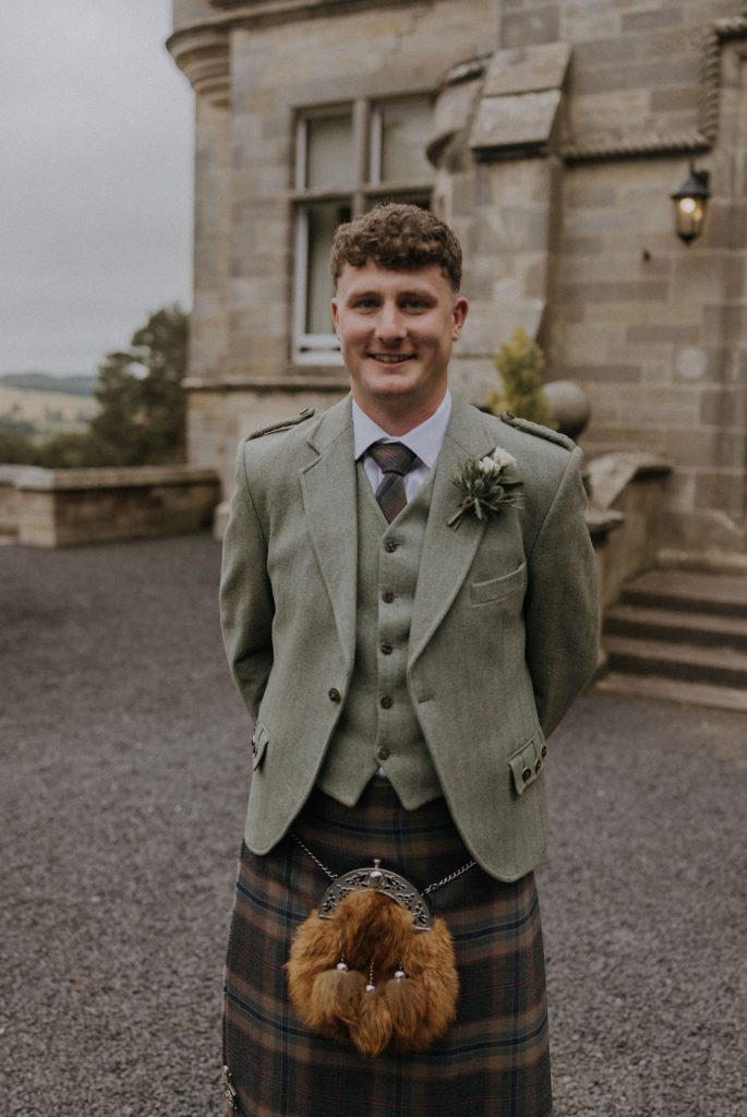 angus-wedding-florist-kinnettles-castle 14 - Fraoch Floral Design Man in a light-green tweed suit and plaid kilt standing outside a stone building, smiling at the camera; sporran hanging from his kilt with a boutonniere on his lapel