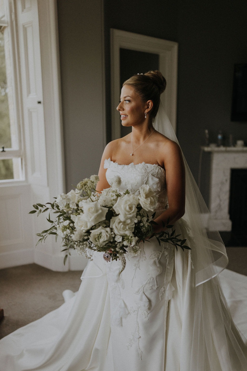 angus-wedding-florist-kinnettles-castle 16 - Fraoch Floral Design Bride in a strapless white wedding gown holding a large bouquet of white roses and greenery, standing by a window.