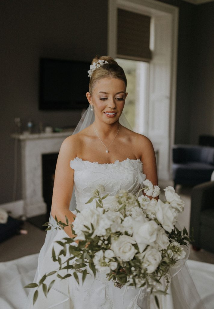 angus-wedding-florist-kinnettles-castle 17 - Fraoch Floral Design Bride in a strapless lace wedding gown holding a lush white bouquet indoors.