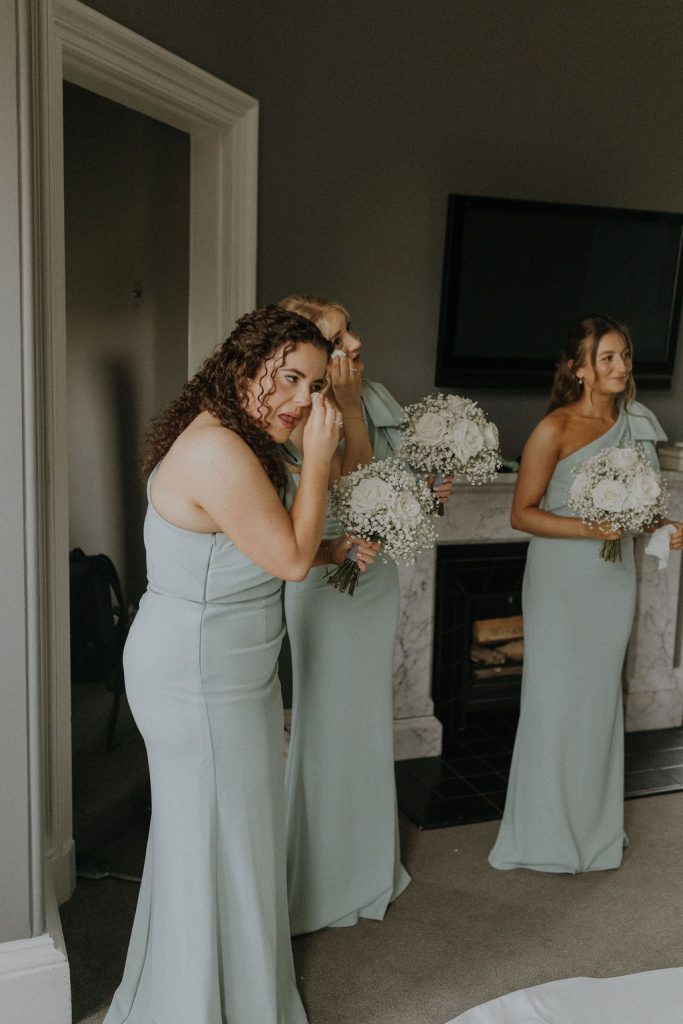 angus-wedding-florist-kinnettles-castle 18 - Fraoch Floral Design Bridesmaids in pale blue gowns holding white bouquet, wiping away tears backstage before a wedding in a living room setting.