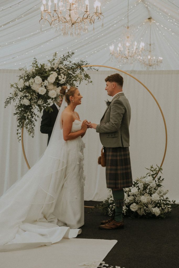 angus-wedding-florist-kinnettles-castle 2 - Fraoch Floral Design Bride and groom share vows under a circular floral arch inside a white tent, chandeliers overhead, wedding gown flowing behind.