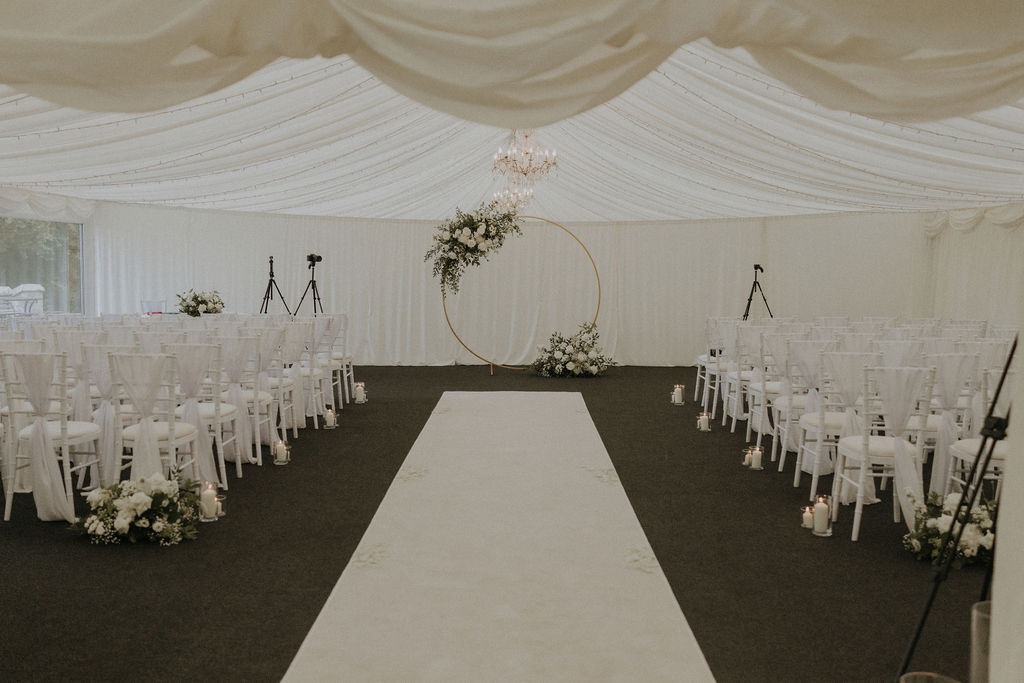 angus-wedding-florist-kinnettles-castle 22 - Fraoch Floral Design White ceremony aisle inside a tent with a circular floral arch at the front and candles along the chairs.