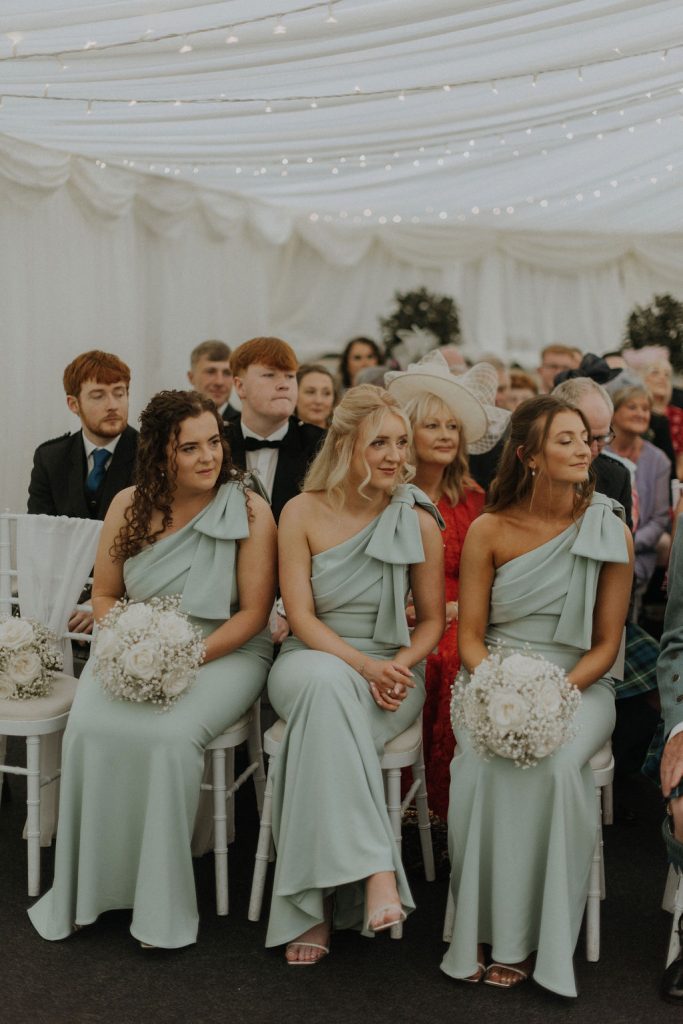 angus-wedding-florist-kinnettles-castle 26 - Fraoch Floral Design Three bridesmaids in pastel teal one-shoulder gowns sit in a wedding venue, each holding white bouquet under twinkling string lights.