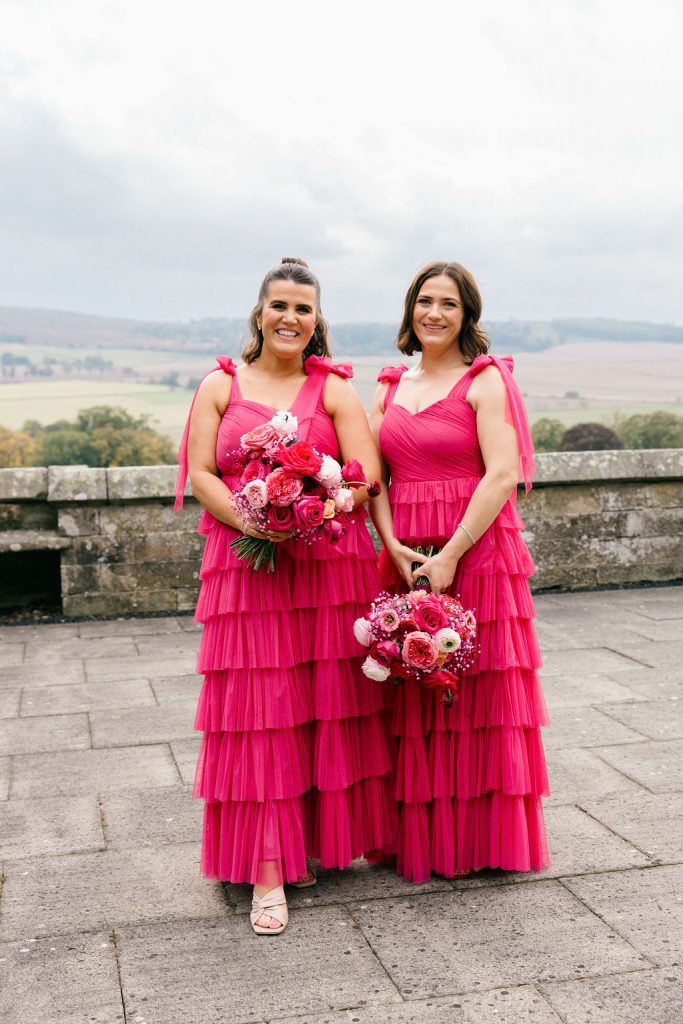 angus-wedding-flowers-florist-fraoch-floral-design 1 - Fraoch Floral Design Two women in matching pink tiered dresses hold bouquets on a stone terrace with a pastoral countryside view behind them.