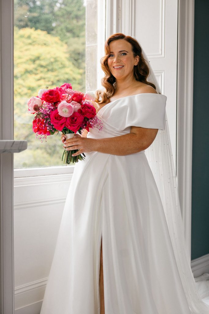 angus-wedding-flowers-florist-fraoch-floral-design 10 - Fraoch Floral Design Bride in an off-shoulder white wedding gown holds a vibrant pink bouquet by a window, smiling at the camera.
