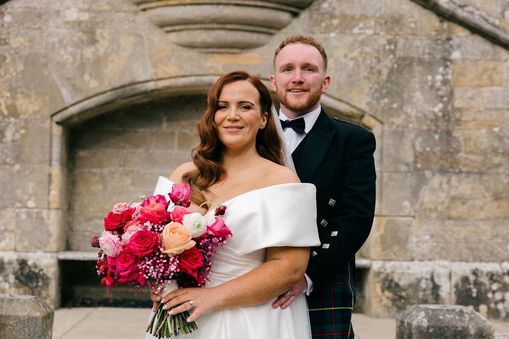 angus-wedding-flowers-florist-fraoch-floral-design 3 - Fraoch Floral Design Bride in a white off-shoulder gown holds a vibrant pink and peach bouquet while standing with a groom in a black tux and tartan kilt before a historic stone archway.