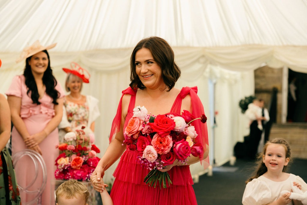angus-wedding-flowers-florist-fraoch-floral-design 4 - Fraoch Floral Design Woman in a bright pink dress holding a large bouquet of pink and red roses, smiling at a festive event with children nearby