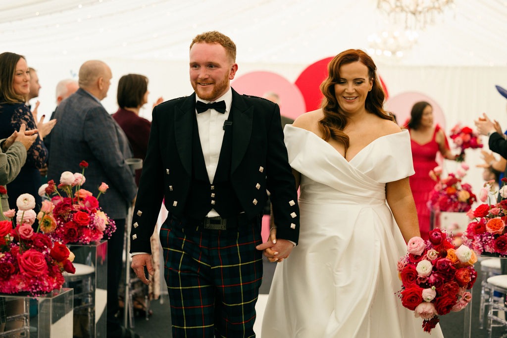 angus-wedding-flowers-florist-fraoch-floral-design 9 - Fraoch Floral Design Bride and groom walk hand in hand down the aisle, smiling, with pink and red flower arrangements around them at their wedding venue.
