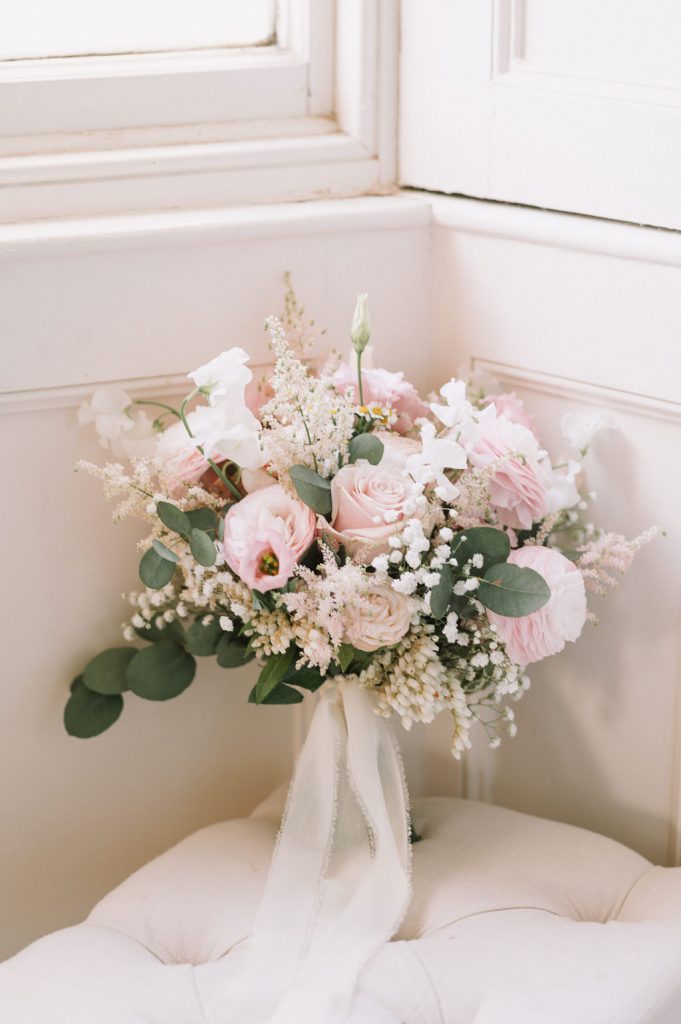 Pastel pink wedding bouquet with roses and baby's breath on a white tufted chair by a window, tied with a white ribbon.