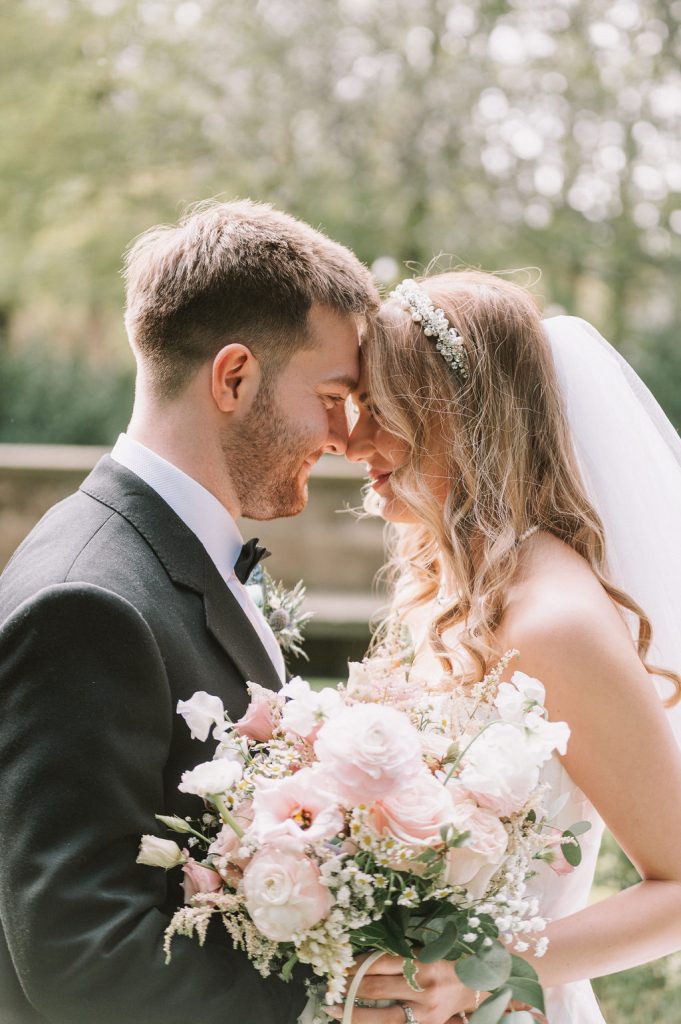 Bride and groom share a kiss, holding a blush pink and white bridal bouquet outdoors.