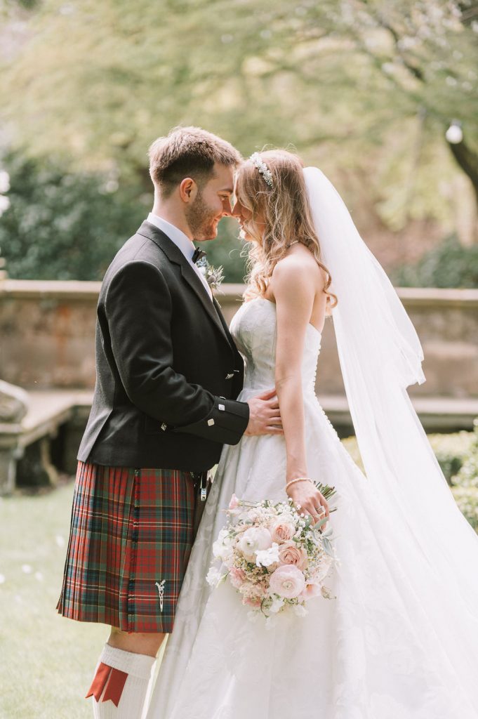 Bride in a white wedding dress and veil leans into a groom in a black jacket and red-green tartan kilt outdoors, holding a blush bouquet.