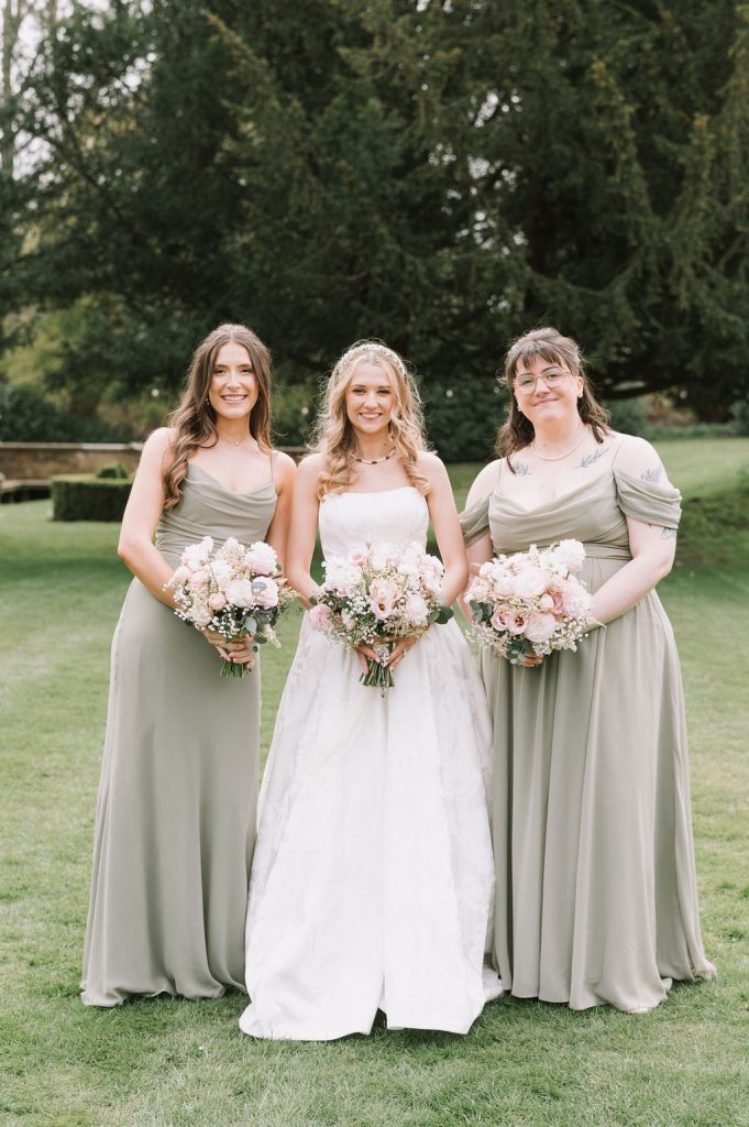 Bride in a white gown stands center with two bridesmaids in sage green dresses, all holding pastel bouquets on a grassy lawn.